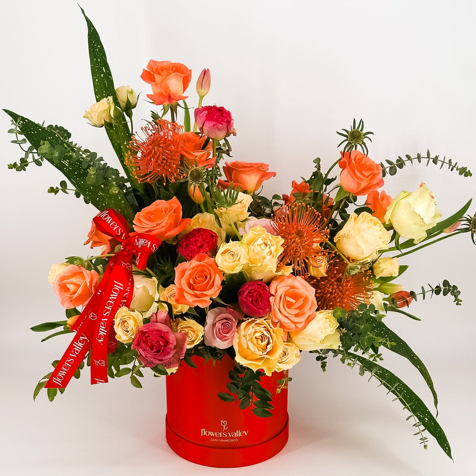 Sunset-colored flower arrangement in a red Flowers Valley box with ribbon, featuring roses, proteas, and greenery.