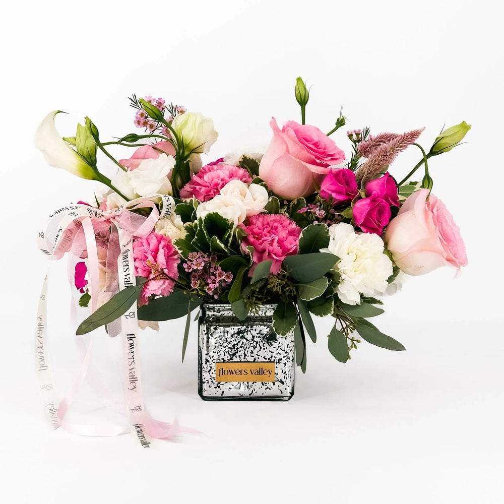Blushing Grace bouquet featuring blush roses, pink spray roses, carnations, and lisianthus in a mirrored vase with Flowers Valley ribbon.