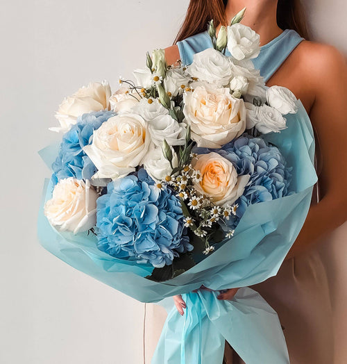 Blue Joy bouquet with blue hydrangeas, white roses, and peach ranunculus wrapped in blue paper