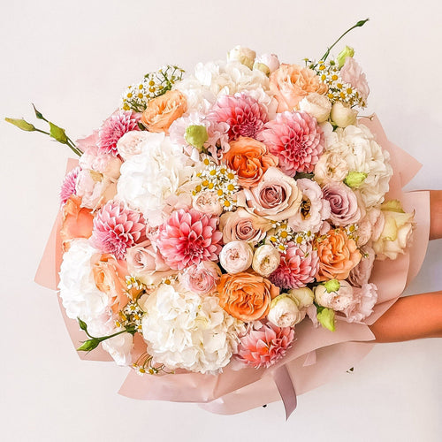 Hand-held bouquet featuring blush dahlias, white hydrangeas, peach roses, lisianthus, and chamomile, wrapped in soft pink paper—fresh, garden-style design.