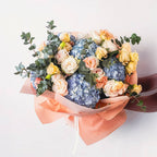Bouquet of peach roses, blue hydrangeas, and greenery wrapped in warm peach-toned paper—fresh and cheerful.