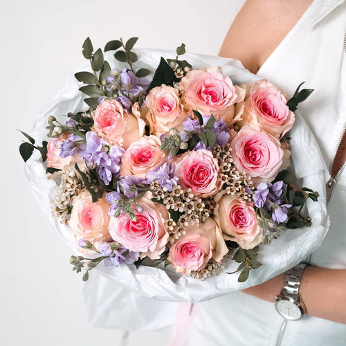 Pink roses with lavender matthiola and white waxflower in a wrapped bouquet.