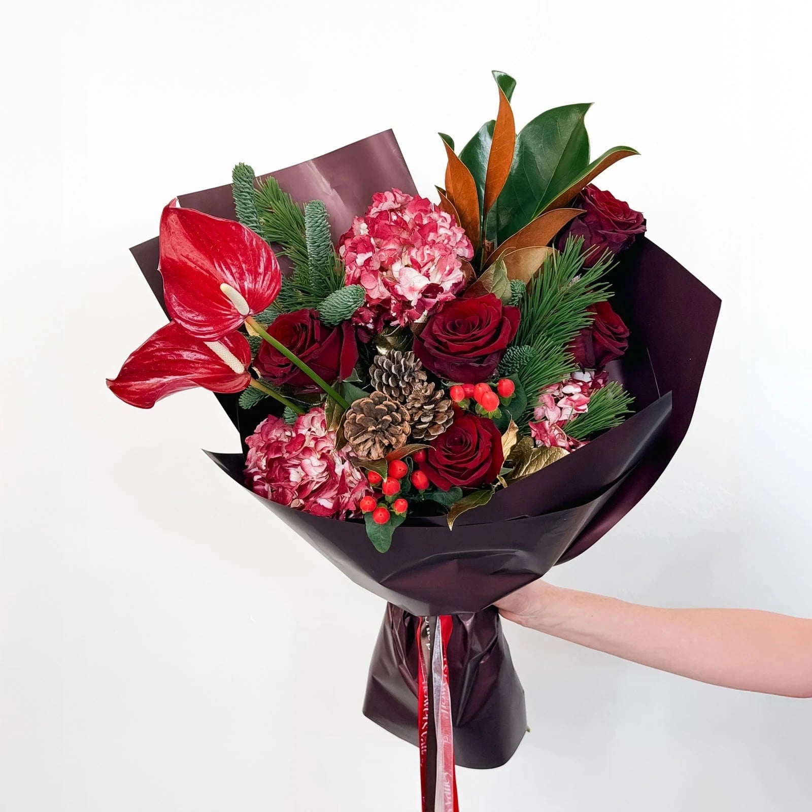 Standard size Crimson Majesty Bouquet with red roses, hydrangeas, protea, and holiday greenery in burgundy wrap