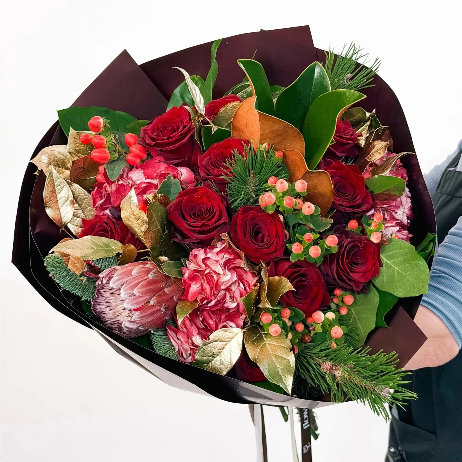 Crimson Majesty Bouquet with red roses, hydrangeas, protea, and holiday greenery in burgundy wrap