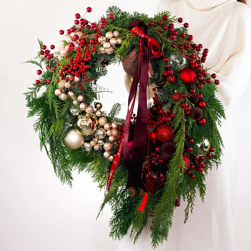Full Crimson Noel Wreath with ornaments, berries, and signature ribbons held by model in white sweater