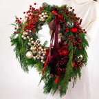Full Crimson Noel Wreath with ornaments, berries, and signature ribbons held by model in white sweater