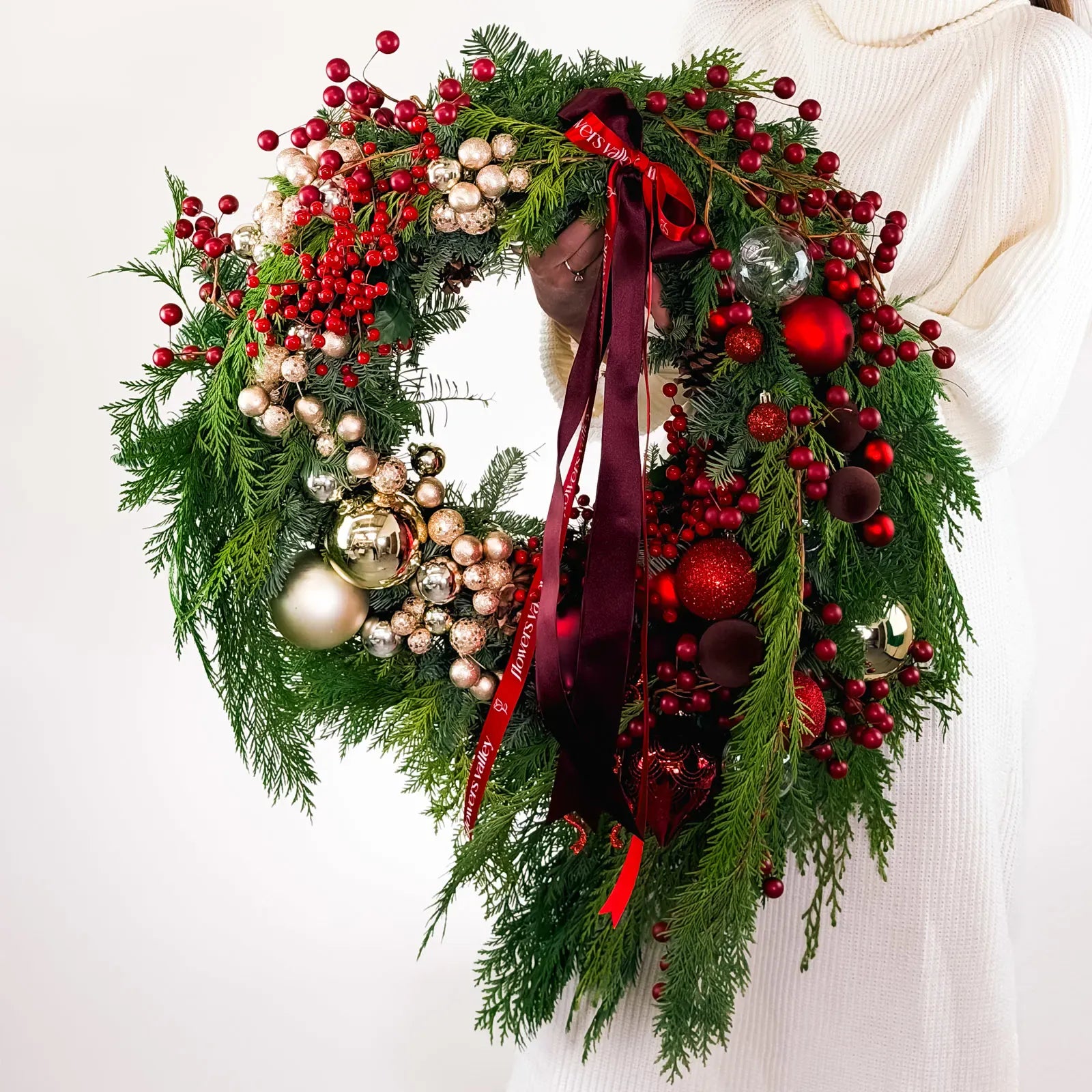 Full Crimson Noel Wreath with ornaments, berries, and signature ribbons held by model in white sweater