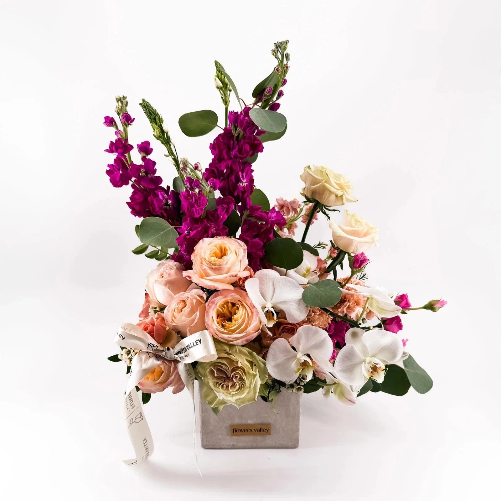 Side view of Majestic Allure premium bouquet with peach roses, magenta stock flowers, and white orchids arranged in a designer vase.