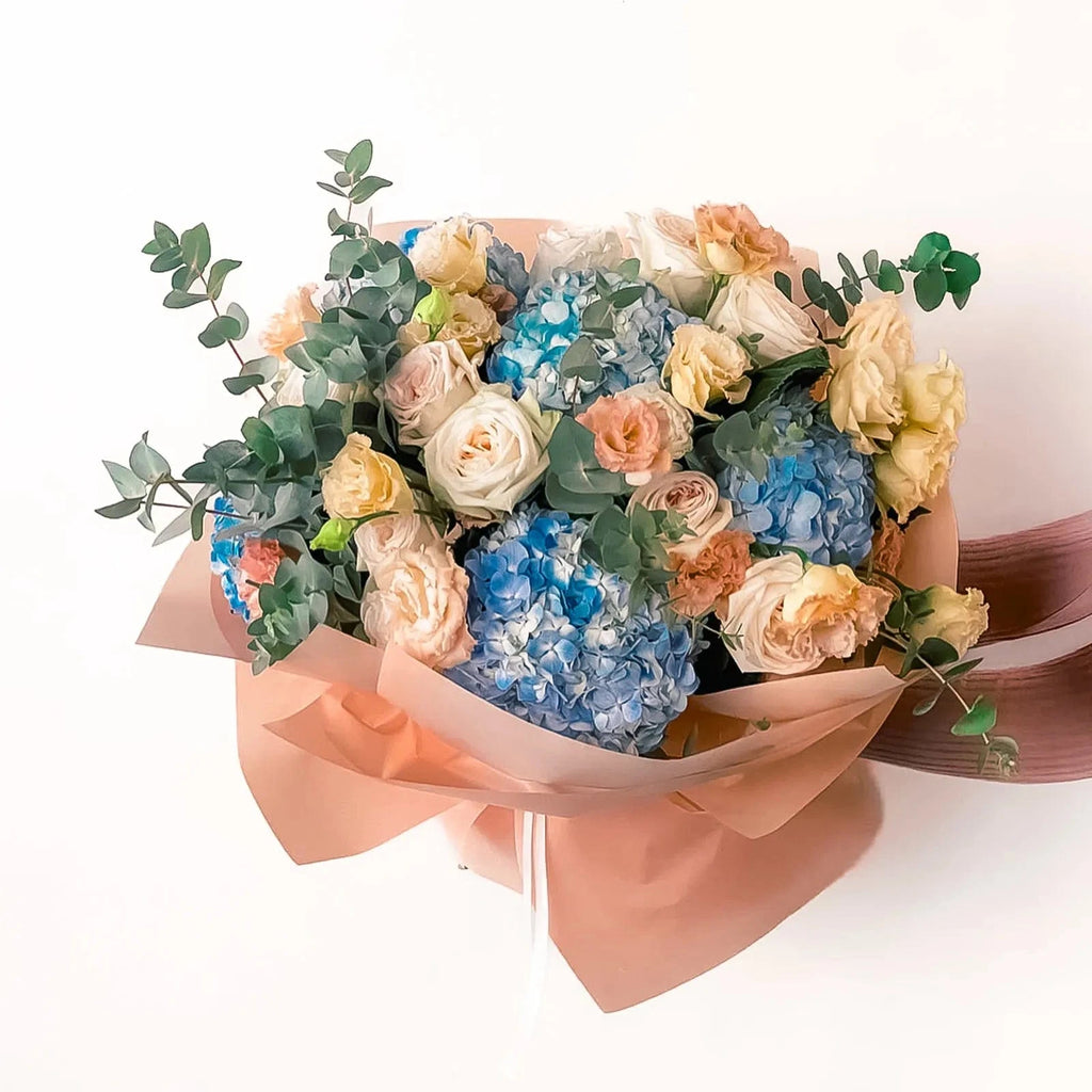 Peach Garden Bouquet in standard size featuring peach roses, blue hydrangeas, and yellow spray roses wrapped in warm-toned paper.