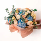 Peach Garden Bouquet in standard size featuring peach roses, blue hydrangeas, and yellow spray roses wrapped in warm-toned paper.