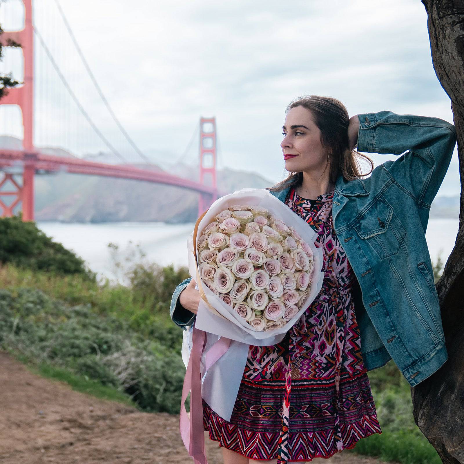 Woman holding a Luxury Quicksand Roses bouquet near the Golden Gate Bridge, showcasing soft blush-beige roses by Flowers Valley.