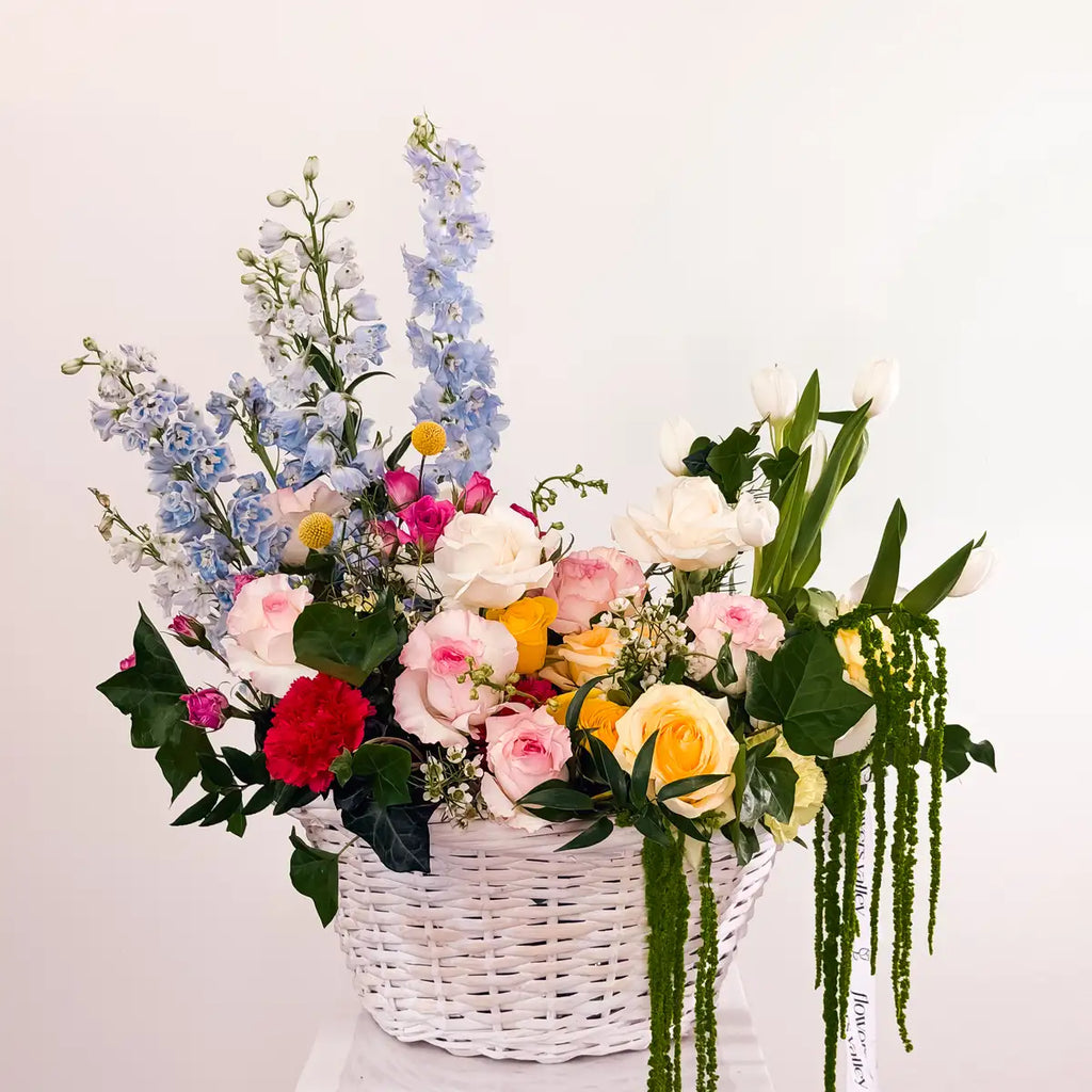 Garden Bloom Flower Basket with blue delphinium, pink garden roses, white tulips, and trailing amaranthus in a white wicker basket by Flowers Valley