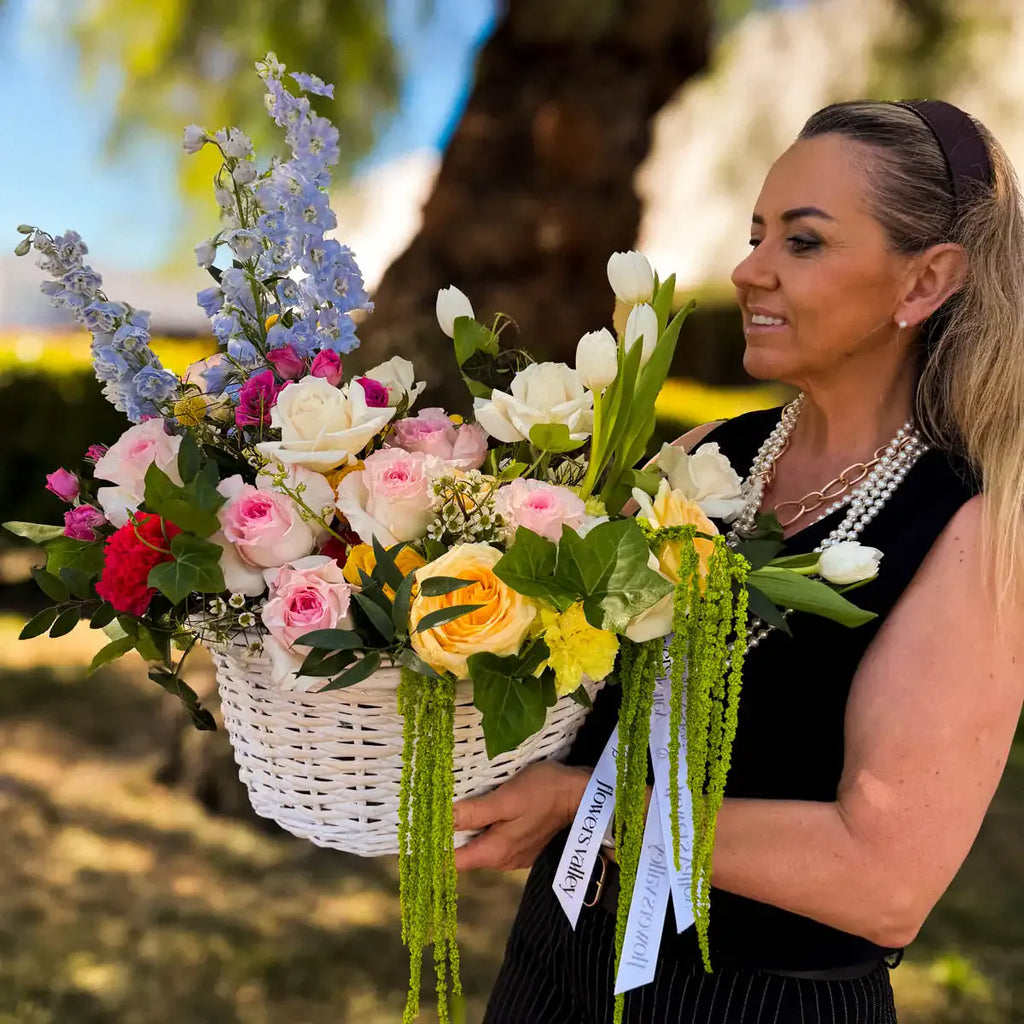 Woman holding Garden Bloom Flower Basket outdoors, colorful fresh flower arrangement with delphinium, roses, and tulips in a white wicker basket