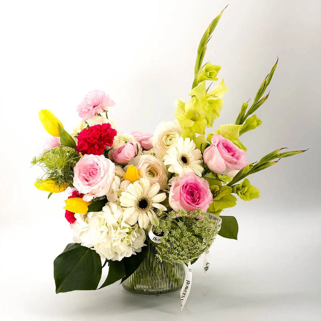 Garden Waltz vase arrangement with pink garden roses, ranunculus, white hydrangea, yellow tulips, and tall green gladiolus in a ribbed glass vase from Flowers Valley