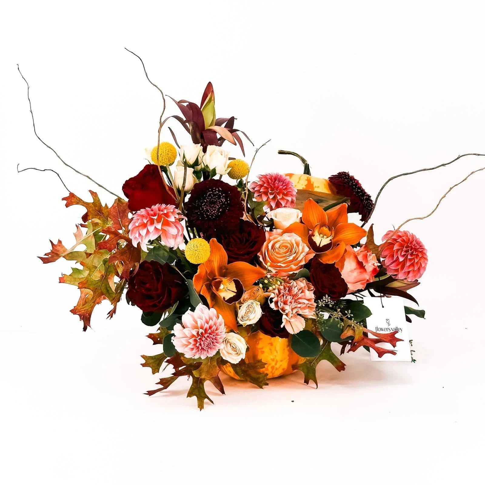 Side angle of Harvest Majesty Pumpkin Arrangement with burgundy, peach, and golden blooms
