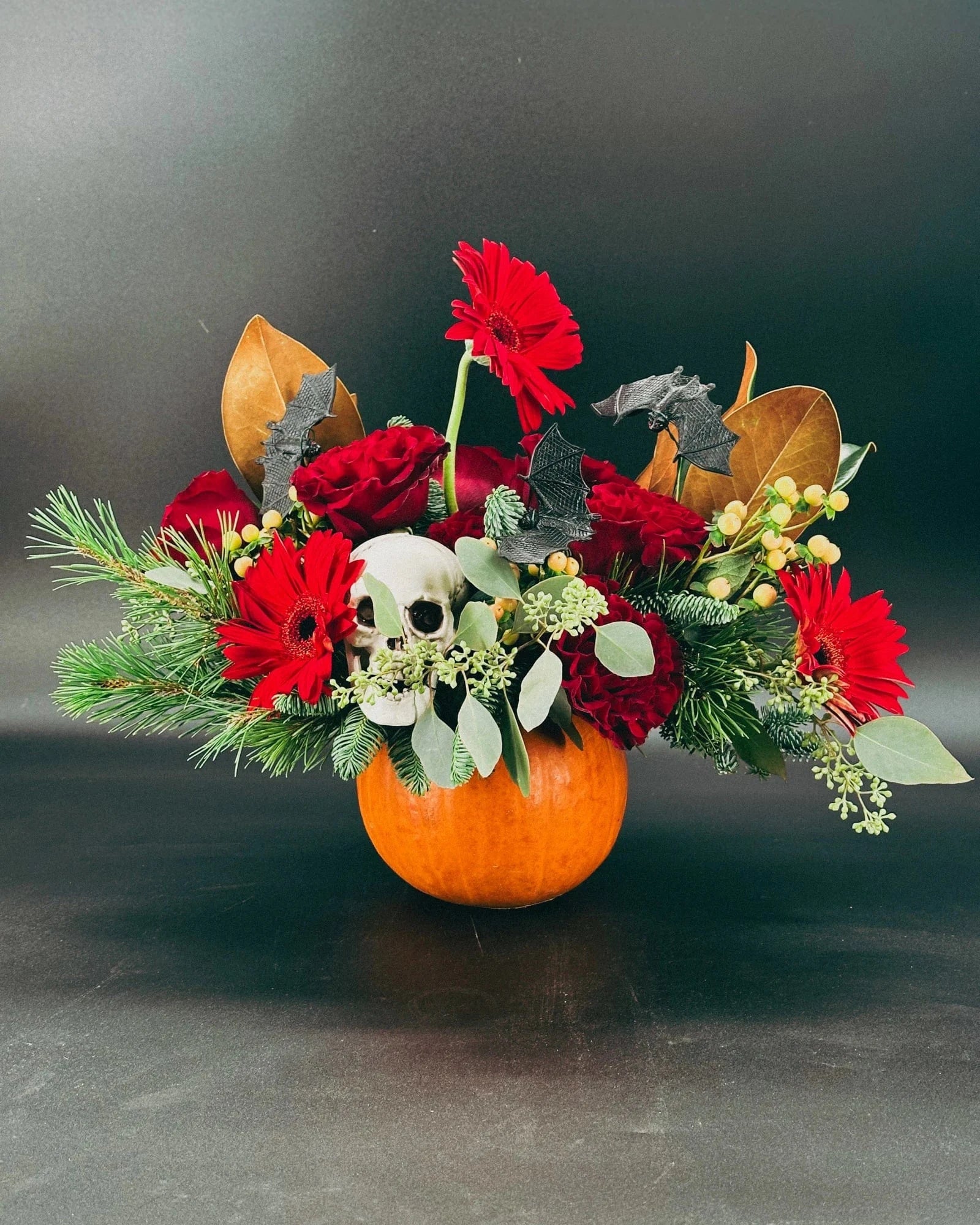 Haunted Elegance Pumpkin Arrangement with skull, bats, and red flowers against dark background