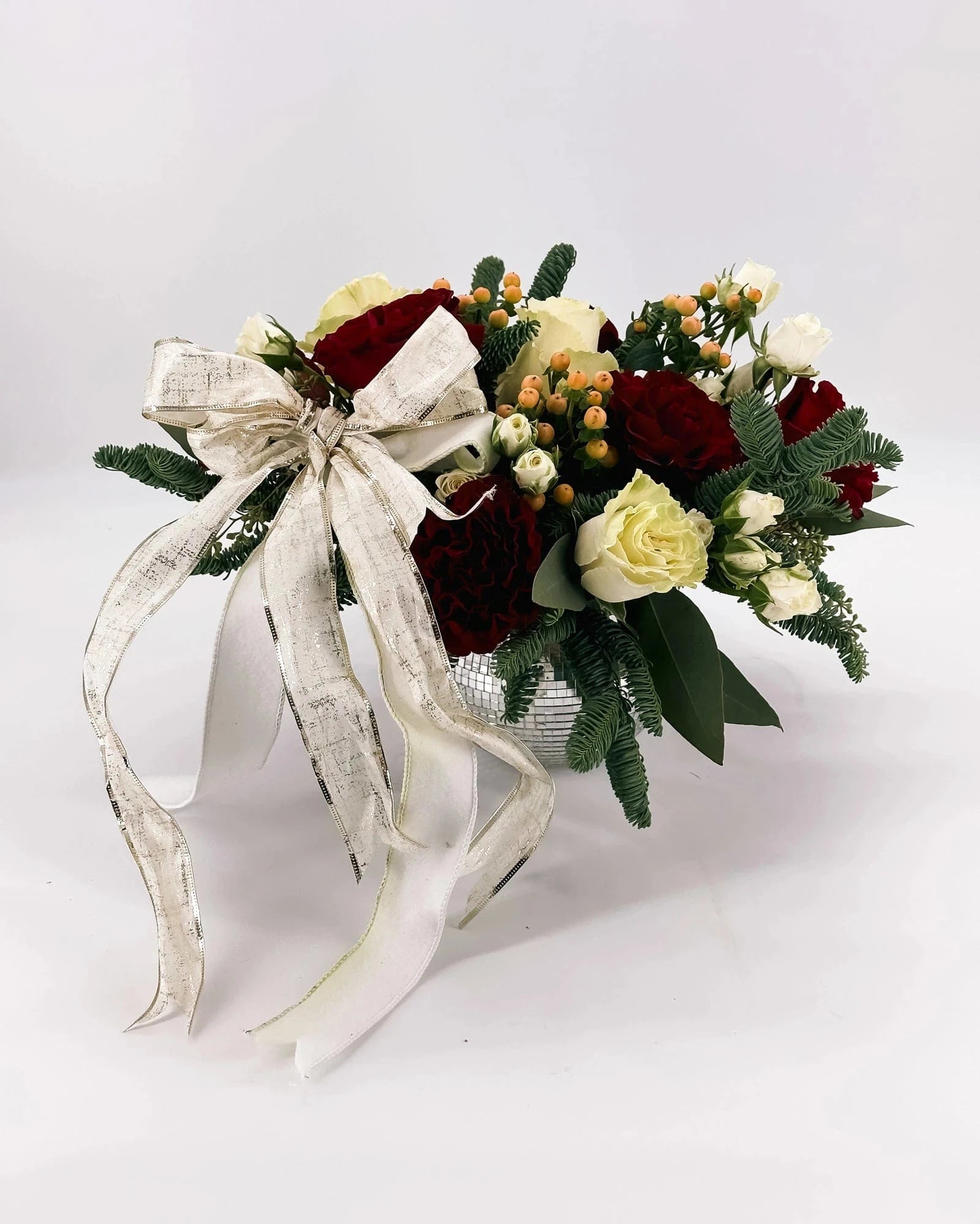 Angled view of Holiday Enchantment Arrangement in silver vase with holiday blooms and gold ribbon