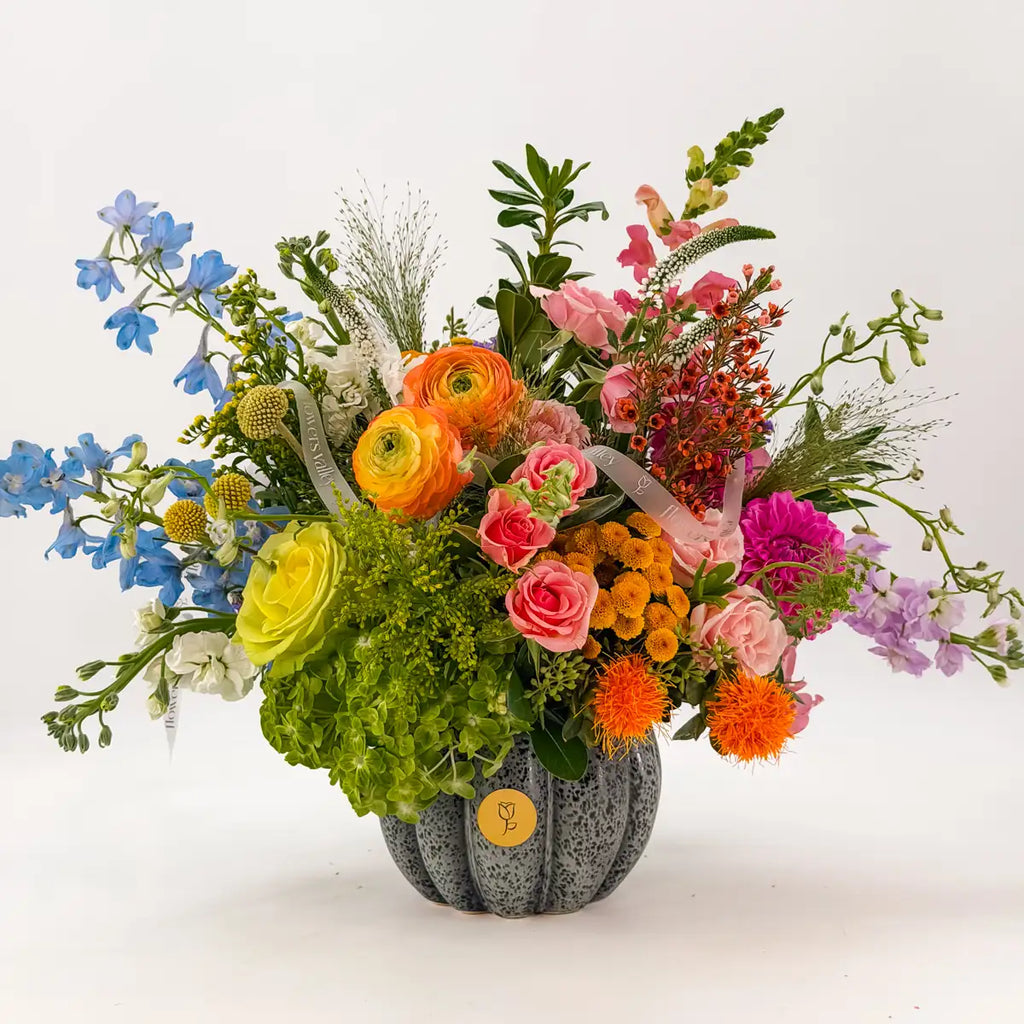 Indigo Harvest fall flower arrangement with blue delphinium, orange ranunculus, pink roses and hydrangea in a grey ceramic pumpkin vase by Flowers Valley