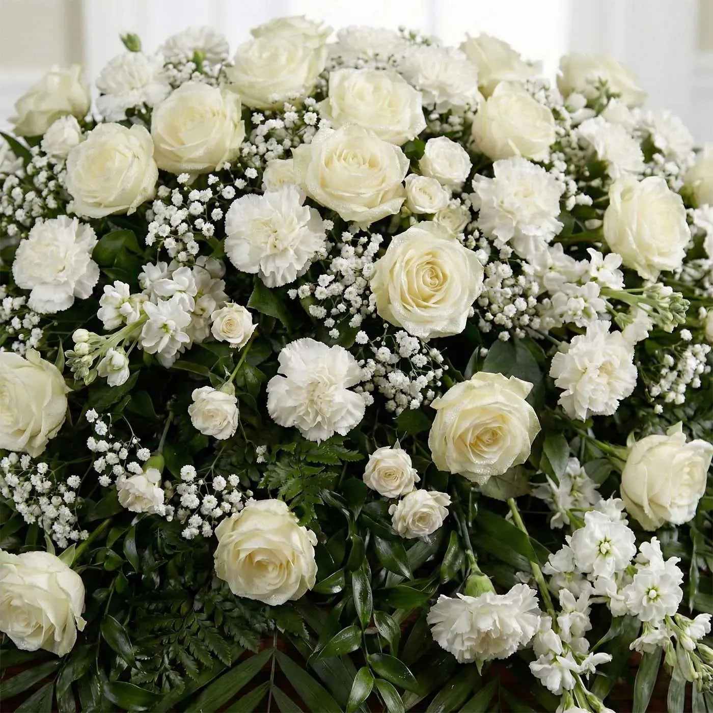 Close-up of white casket arrangement with ivory roses, white carnations, baby’s breath, and greenery