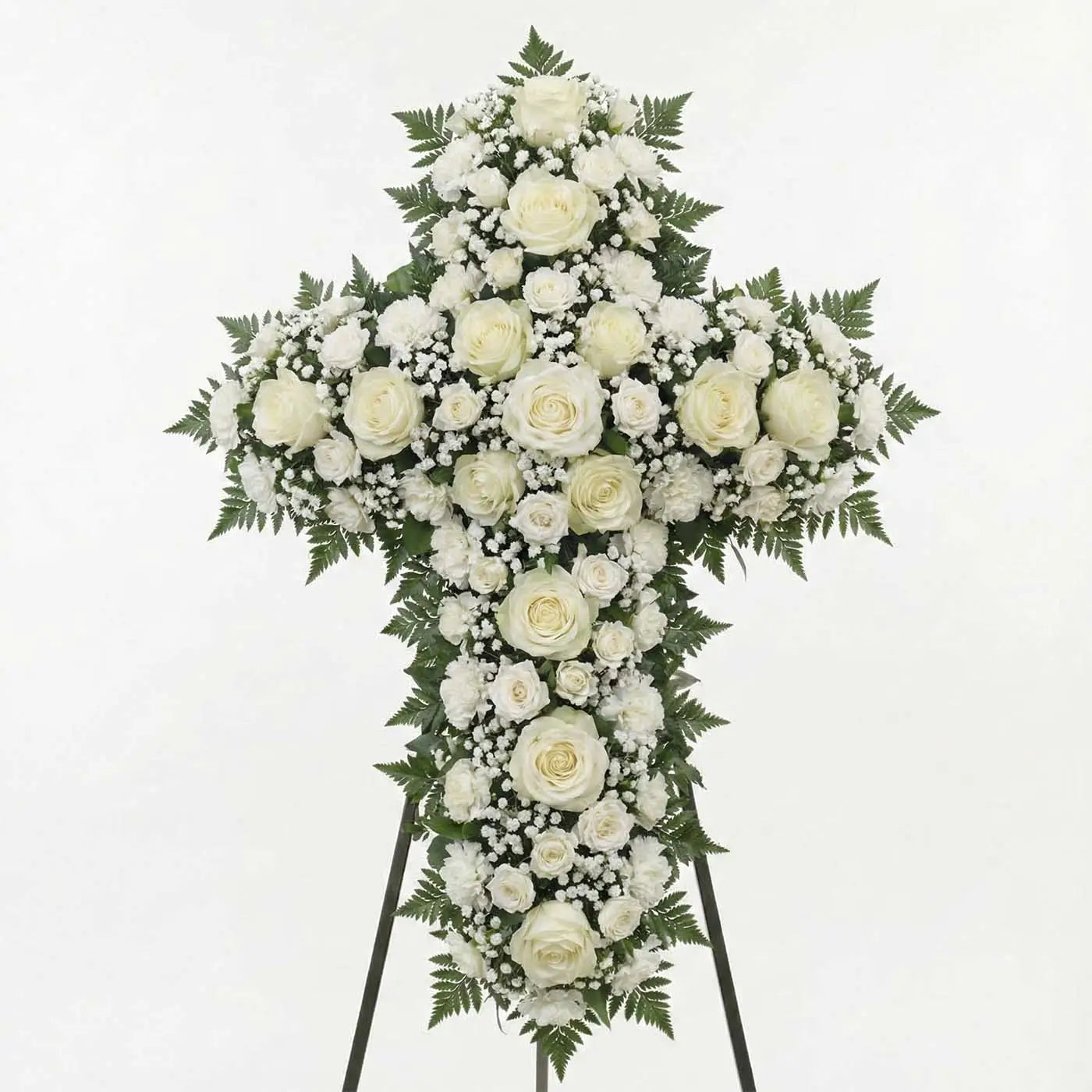 White standing floral cross with ivory roses, white carnations, baby’s breath, and fern greenery