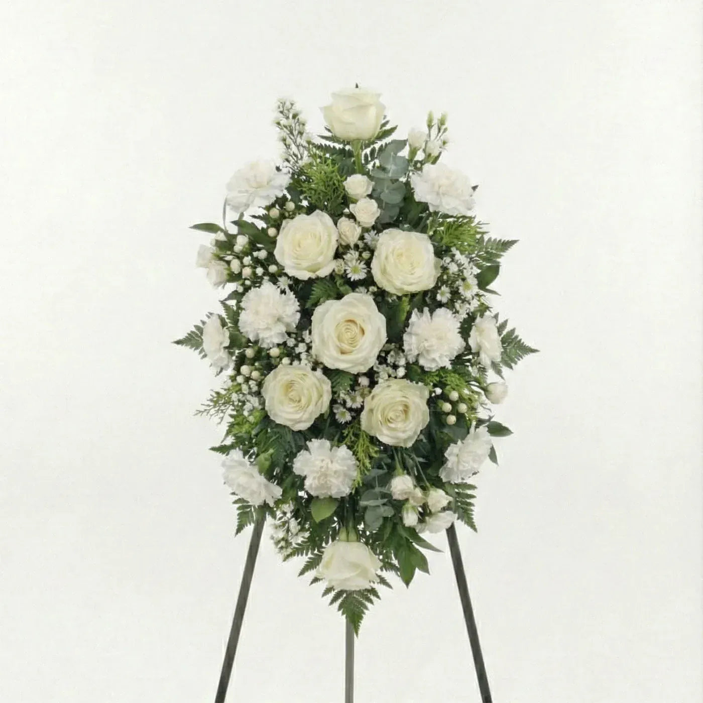 Classic white standing funeral spray with ivory roses, white carnations, baby’s breath, and fern greenery on an easel