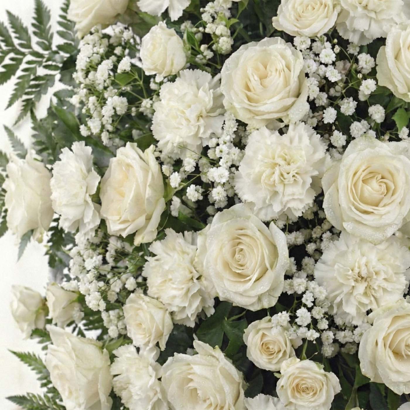 Close-up of standing funeral spray featuring ivory roses, white carnations, baby’s breath, and fern greenery