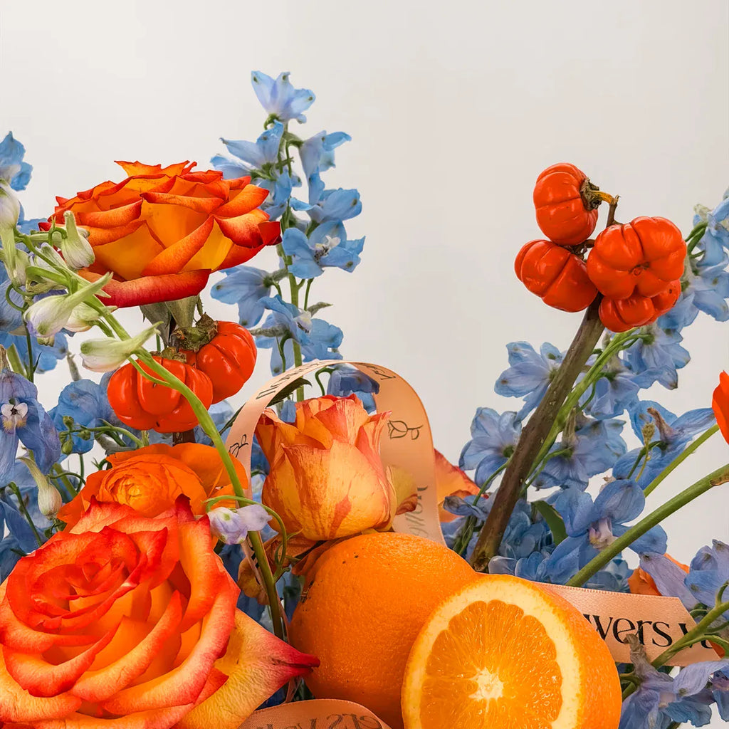 Close-up of orange Free Spirit roses, blue delphinium, pumpkin-on-a-stick and sliced orange in the Libra zodiac arrangement