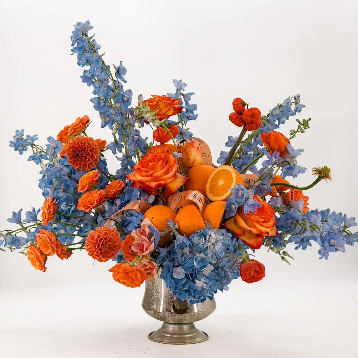 Libra zodiac flower arrangement with blue delphinium, hydrangea, orange roses, dahlias and fresh oranges in a vintage silver urn by Flowers Valley
