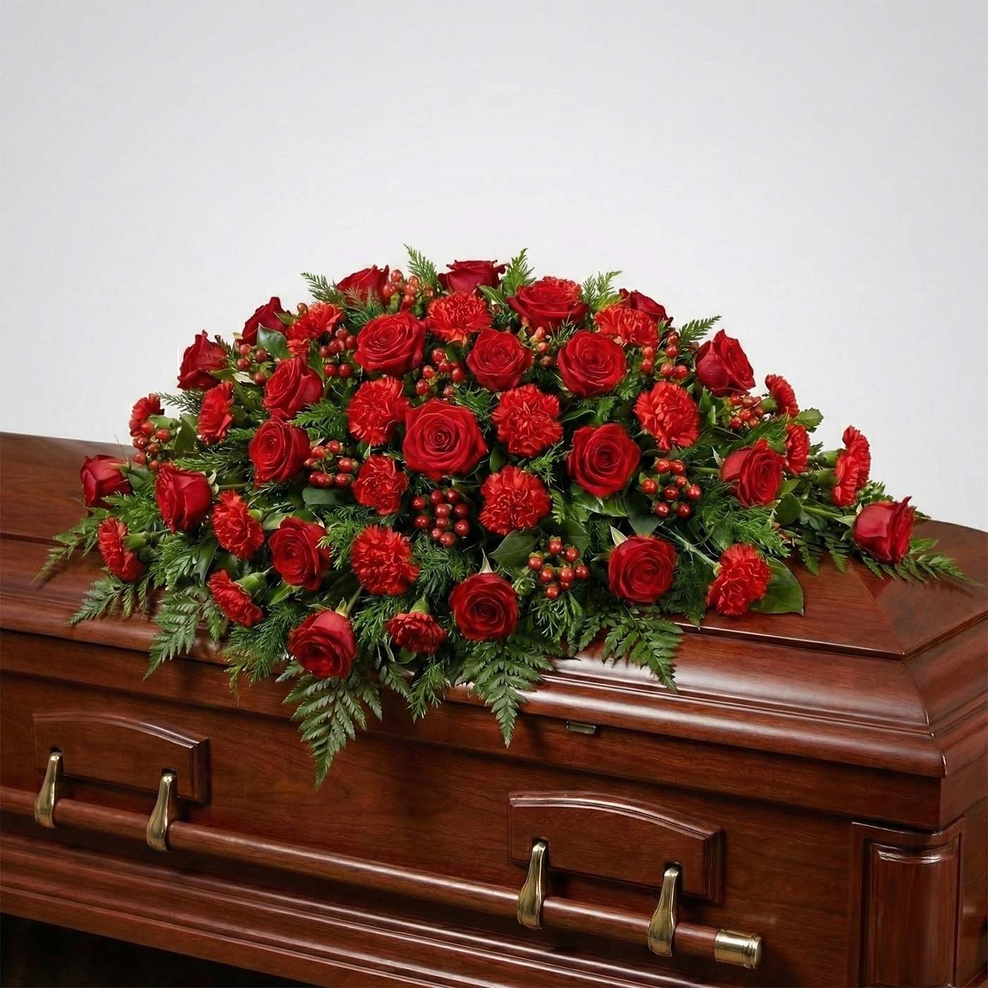 Half casket funeral arrangement with deep red roses, red carnations, hypericum berries, and evergreen fern greenery placed atop a casket.
