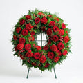 Circular funeral wreath with deep red roses, red carnations, hypericum berries, and evergreen fern greenery displayed on a standing easel.