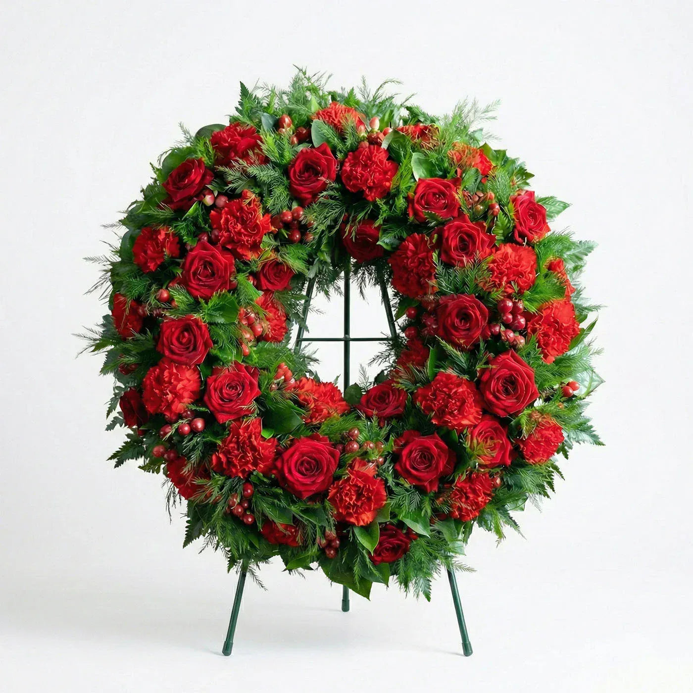 Circular funeral wreath with deep red roses, red carnations, hypericum berries, and evergreen fern greenery displayed on a standing easel.