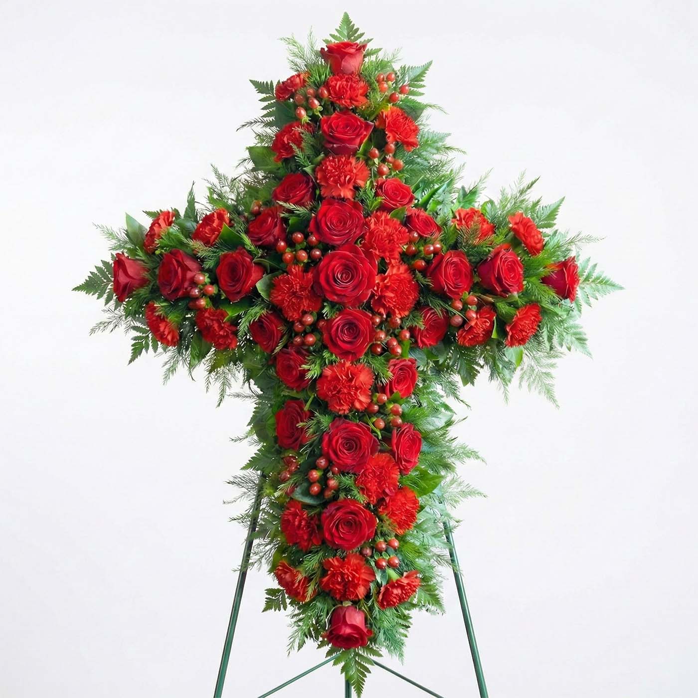 Standing floral cross with deep red roses, red carnations, hypericum berries, and evergreen fern greenery arranged for a funeral service.