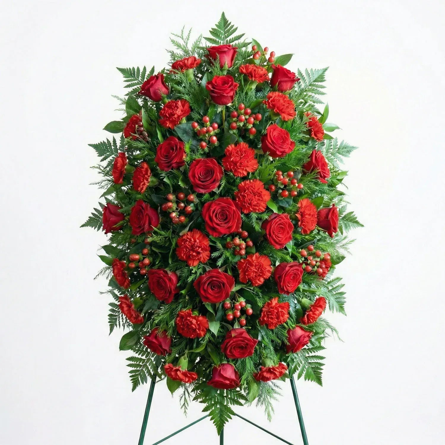 Medium size standing funeral spray with deep red roses, red carnations, hypericum berries, and evergreen fern greenery displayed on an easel.