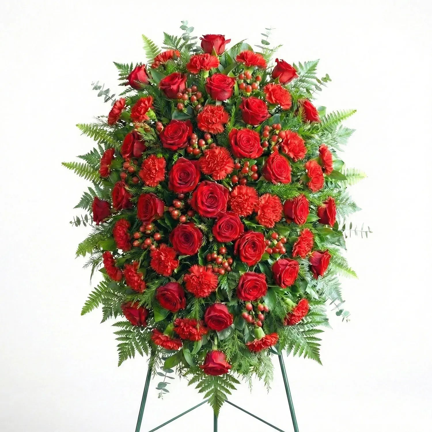 Standing funeral spray with deep red roses, red carnations, hypericum berries, and evergreen fern greenery displayed on an easel.