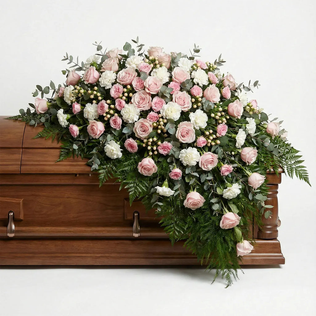 Half casket funeral arrangement with blush roses, white carnations, eucalyptus, and cascading fern greenery placed atop a wooden casket.
