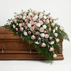 Half casket funeral arrangement with blush roses, white carnations, eucalyptus, and cascading fern greenery placed atop a wooden casket.