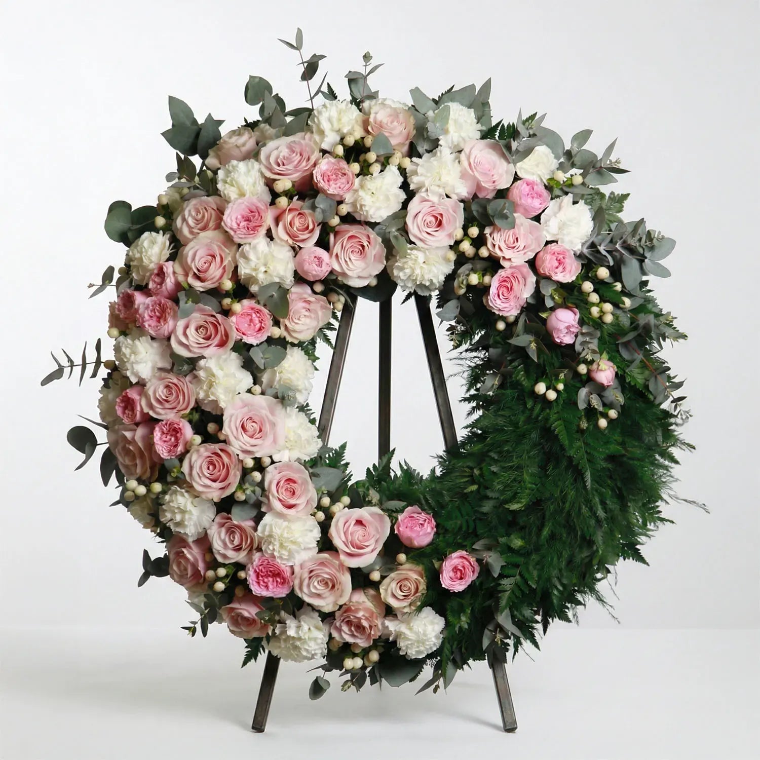 Circular funeral wreath with blush roses, white carnations, eucalyptus, and fern greenery displayed on a standing easel.