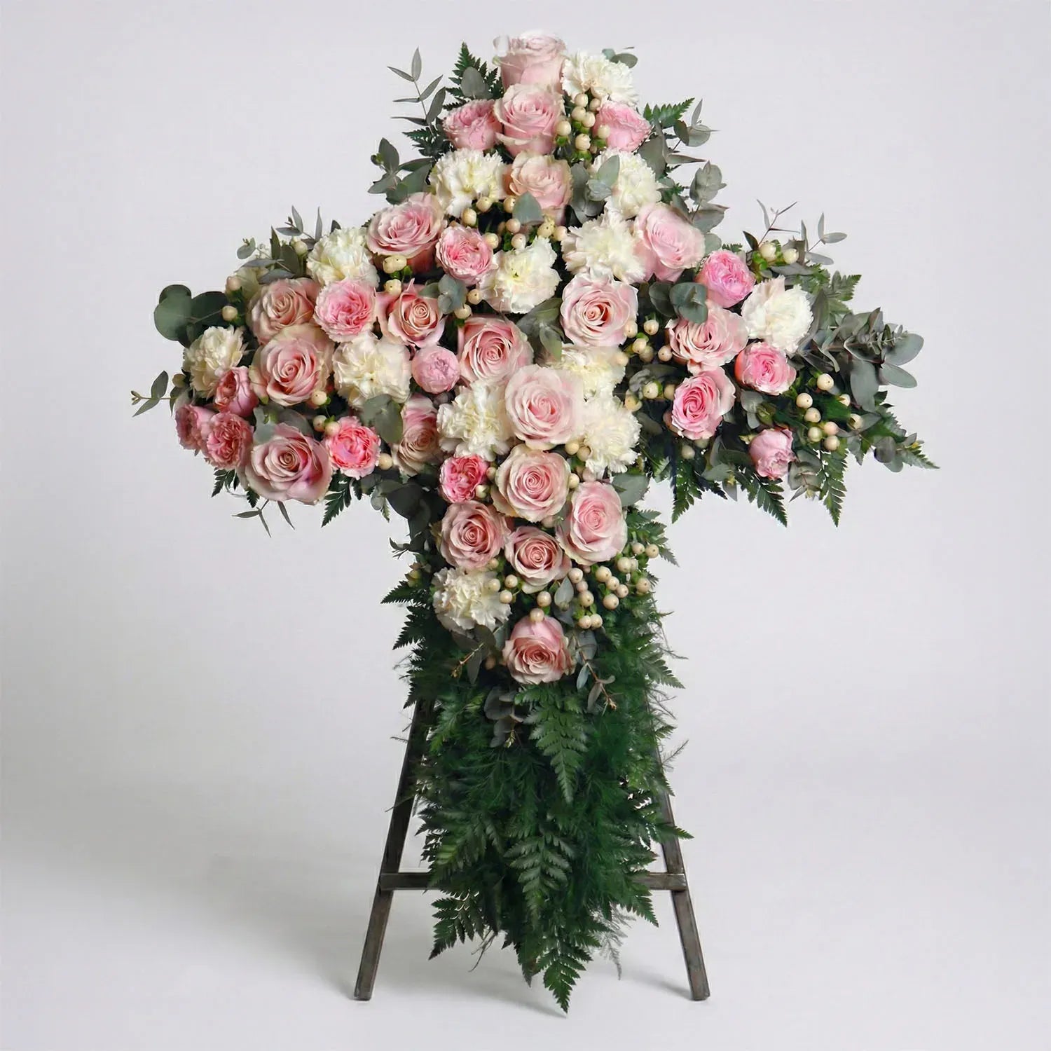 Standing floral cross with blush roses, white carnations, eucalyptus, and fern greenery arranged for a funeral service.