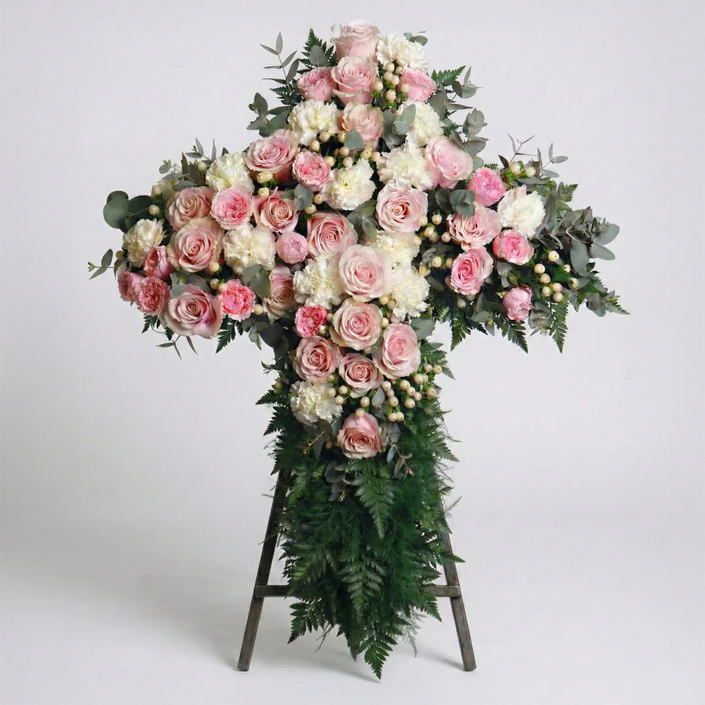 Standing floral cross with blush roses, white carnations, eucalyptus, and fern greenery arranged for a funeral service.