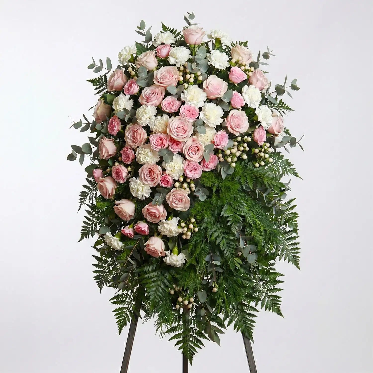 Standing funeral spray with blush roses, white carnations, eucalyptus, and fern greenery arranged in a soft floral gradient on an easel.