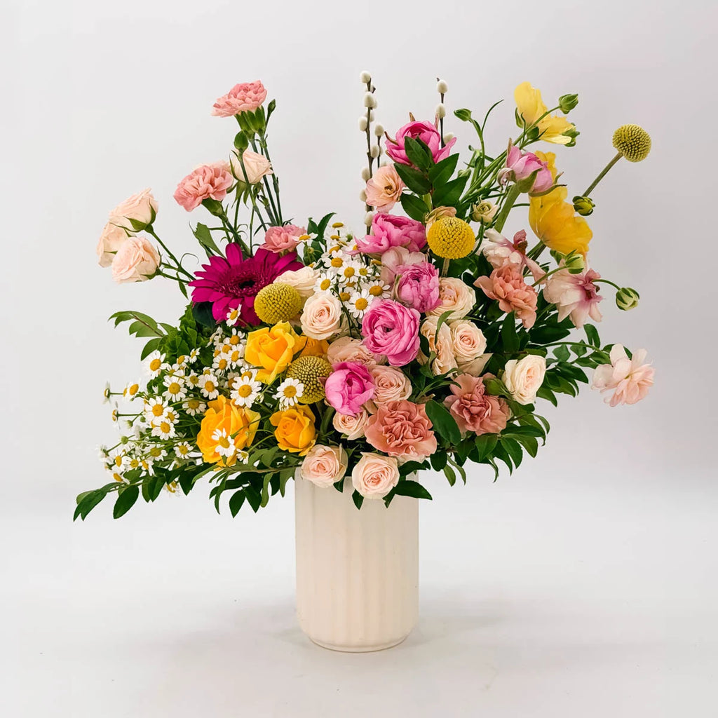 Sunlit Garden vase arrangement featuring pink ranunculus, yellow pon-pon ranunculus, and blush spray roses in a white fluted ceramic vase — Flowers Valley