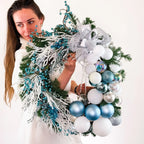 Handcrafted winter wreath with icy blue and white ornaments held by woman in white sweater.