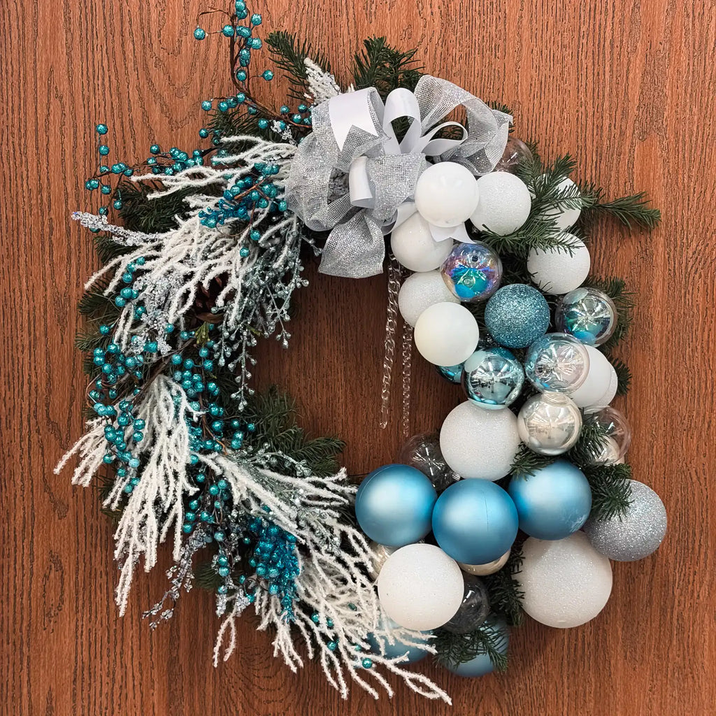 Winter wreath with silver ribbon, frosted foliage, and blue holiday ornaments on wooden background.