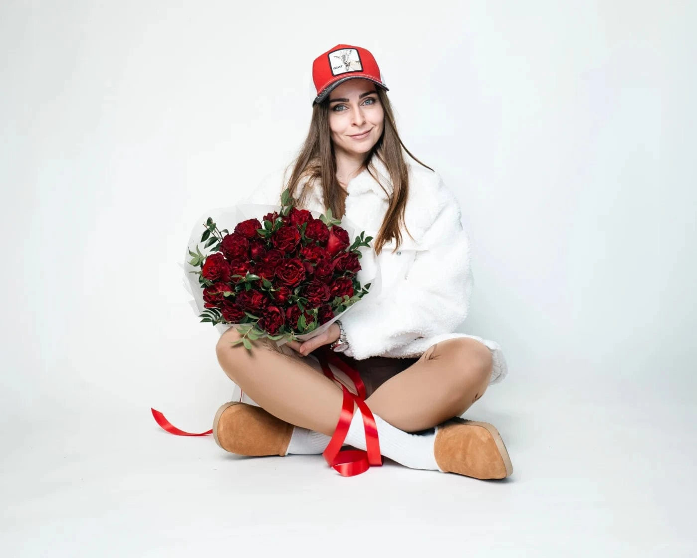 Smiling woman in a red cap sitting cross-legged with a bouquet of red roses and greenery tied with ribbon – Flowers Valley Vacaville flower delivery